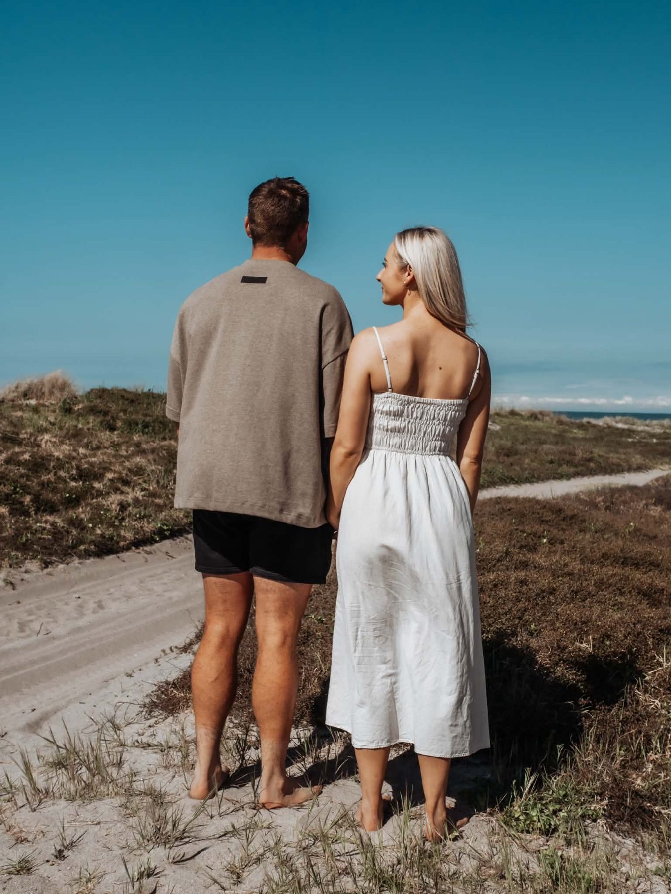 Lacy and Josh standing hand in hand on a sandy coastal track under a blue sky, seen from behind